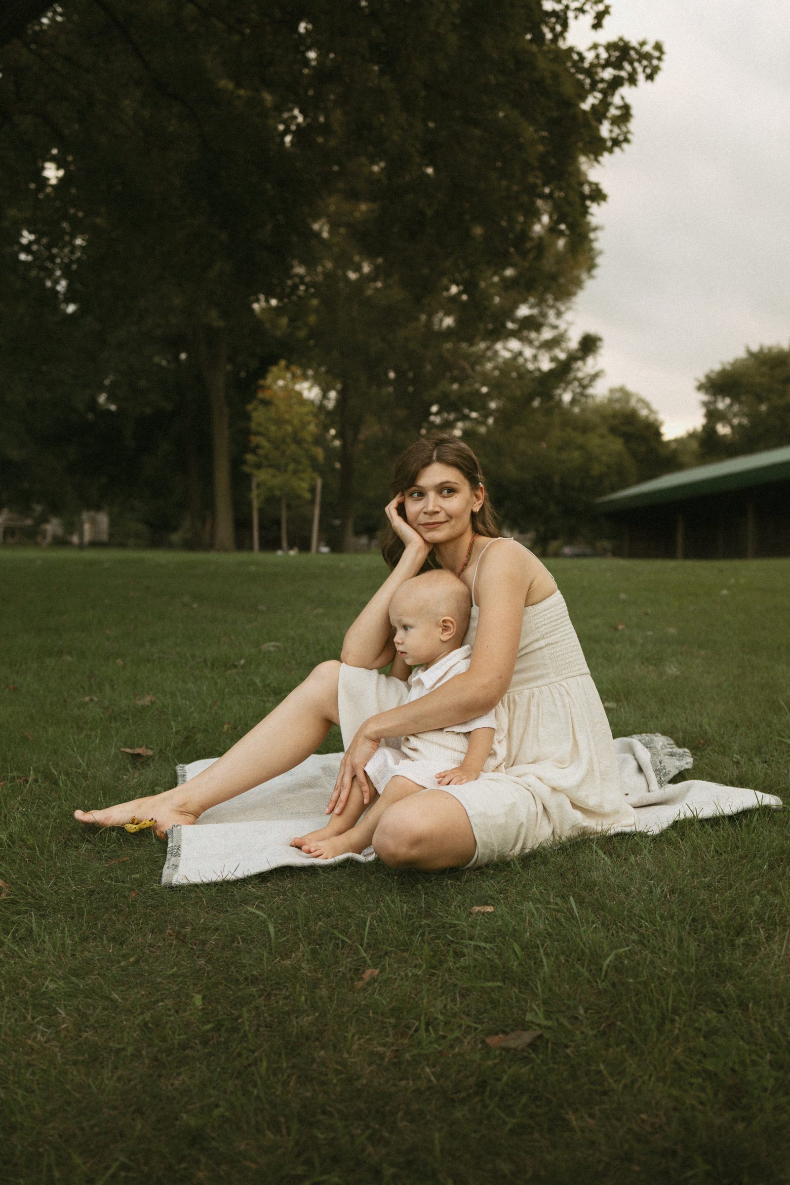 A mother in a cream linen dress sitting barefoot on a blanket in Dickson Park, Cambridge, with her son in her lap
