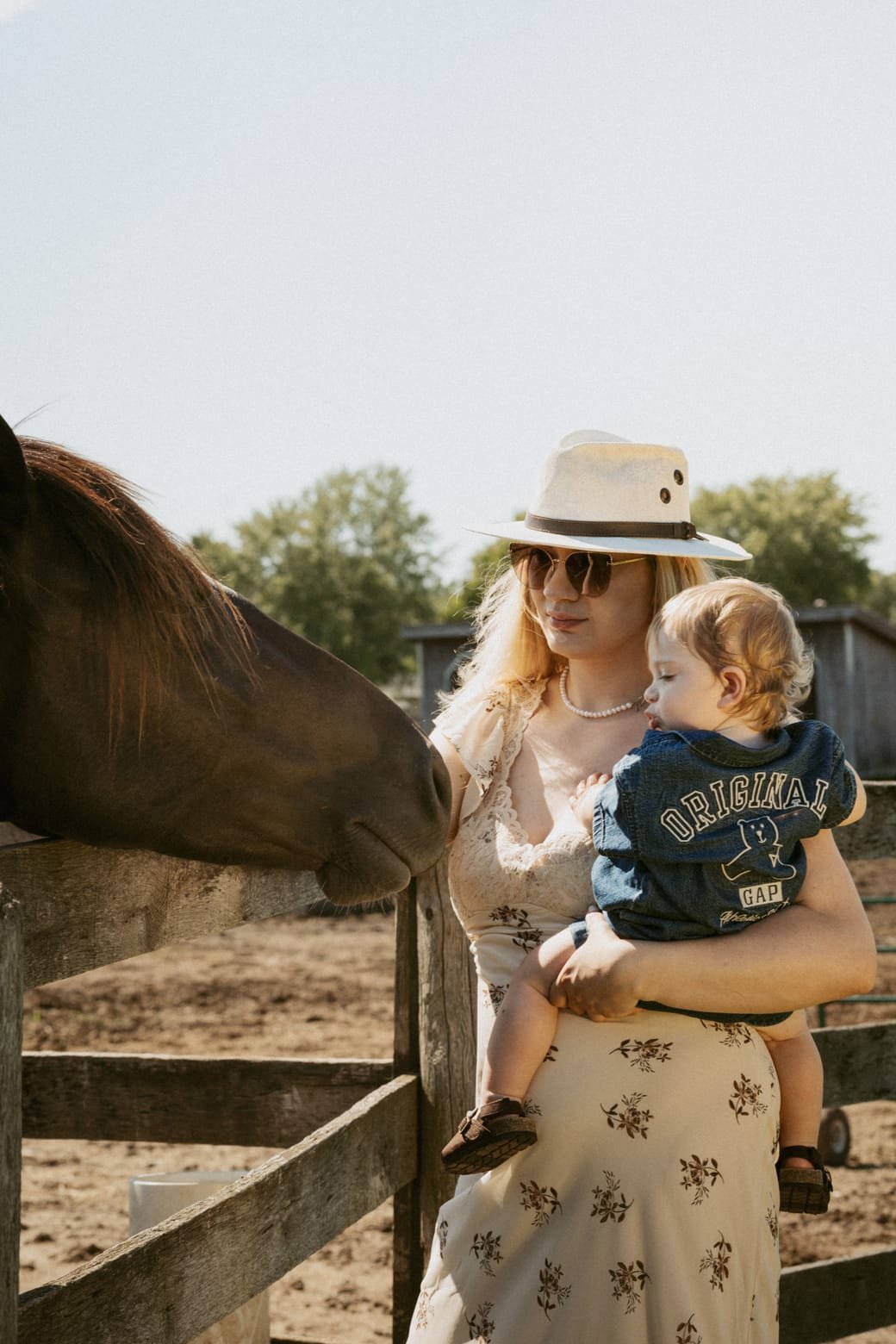 A mother tilting her head down to her son's face while a horse looks on over the fence