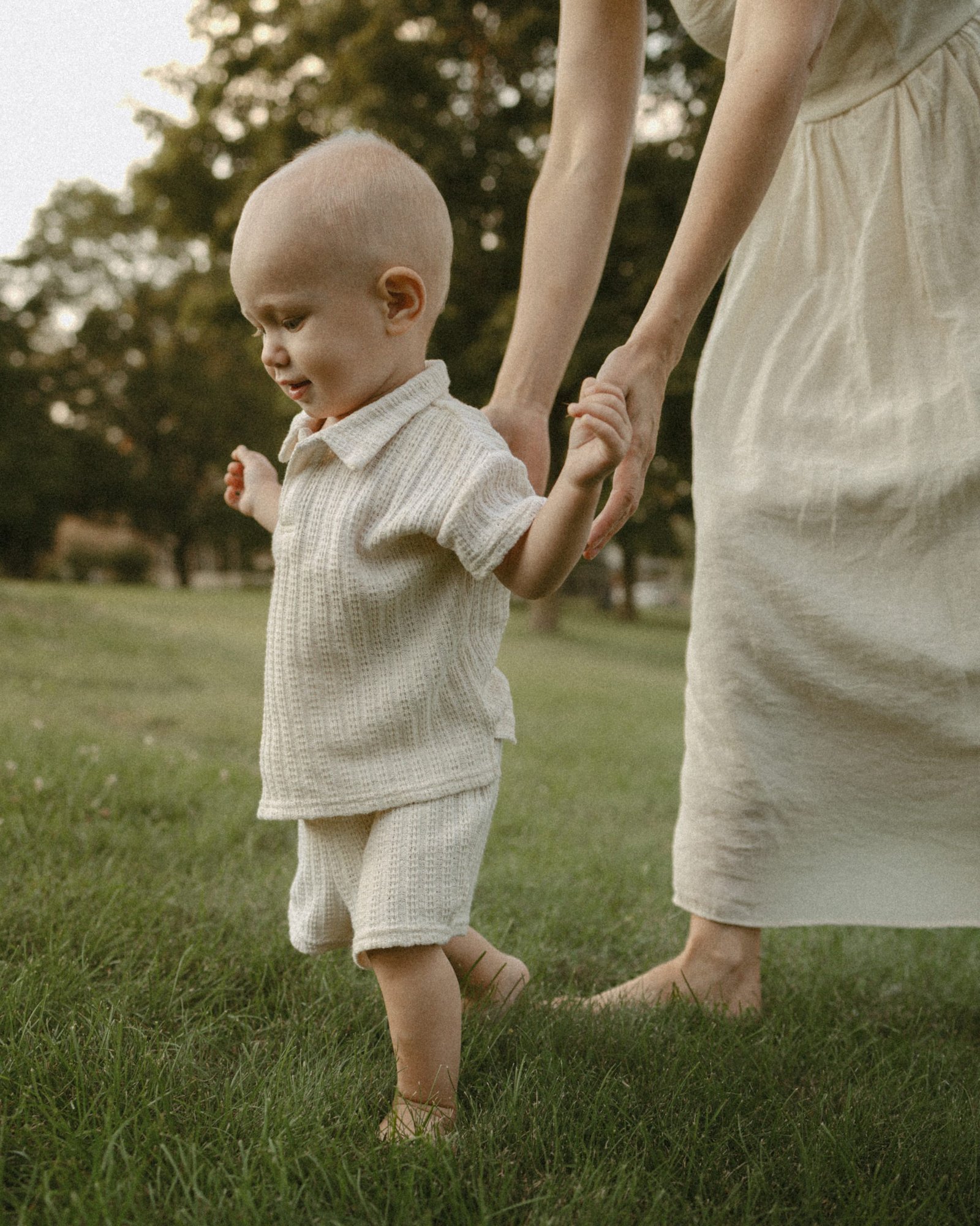 A barefoot baby taking wobbly steps across the grass, holding on to his mother's fingers
