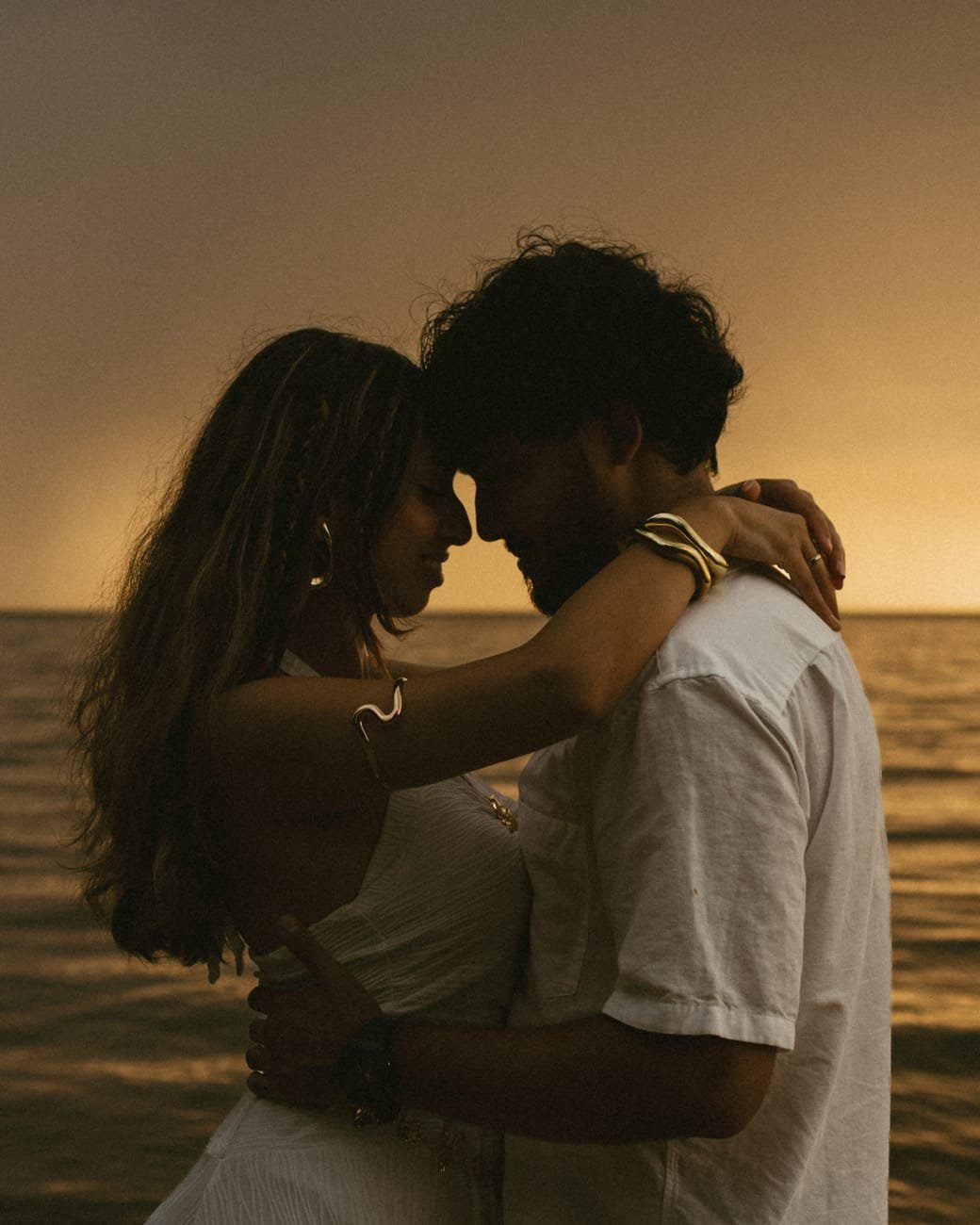 A couple with their foreheads touching at the water's edge in low orange light