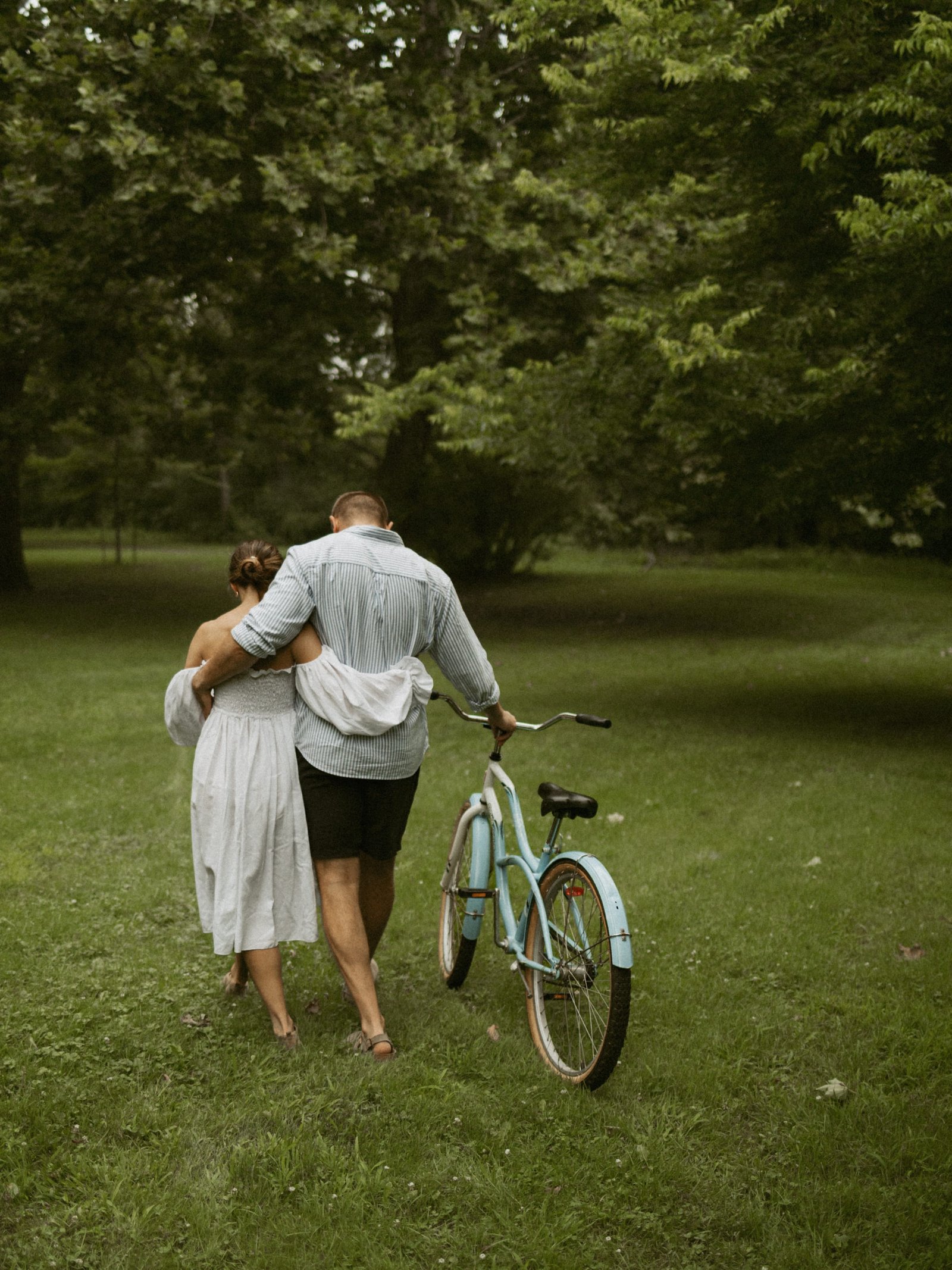 A couple walking hand in hand across a park lawn with a bicycle, seen from behind