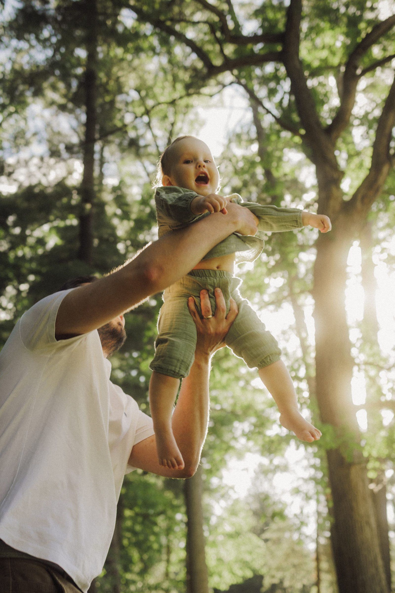 A father lifting his laughing baby overhead beneath tall trees in Victoria Park, Cambridge