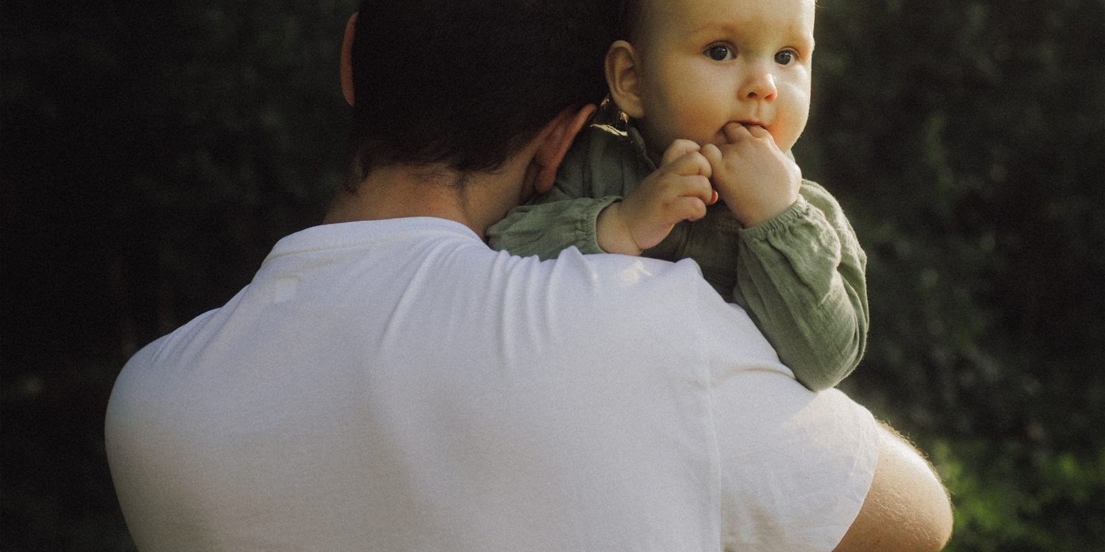 A father carrying his baby son on his shoulder through sunlit woodland, the boy looking back at the camera