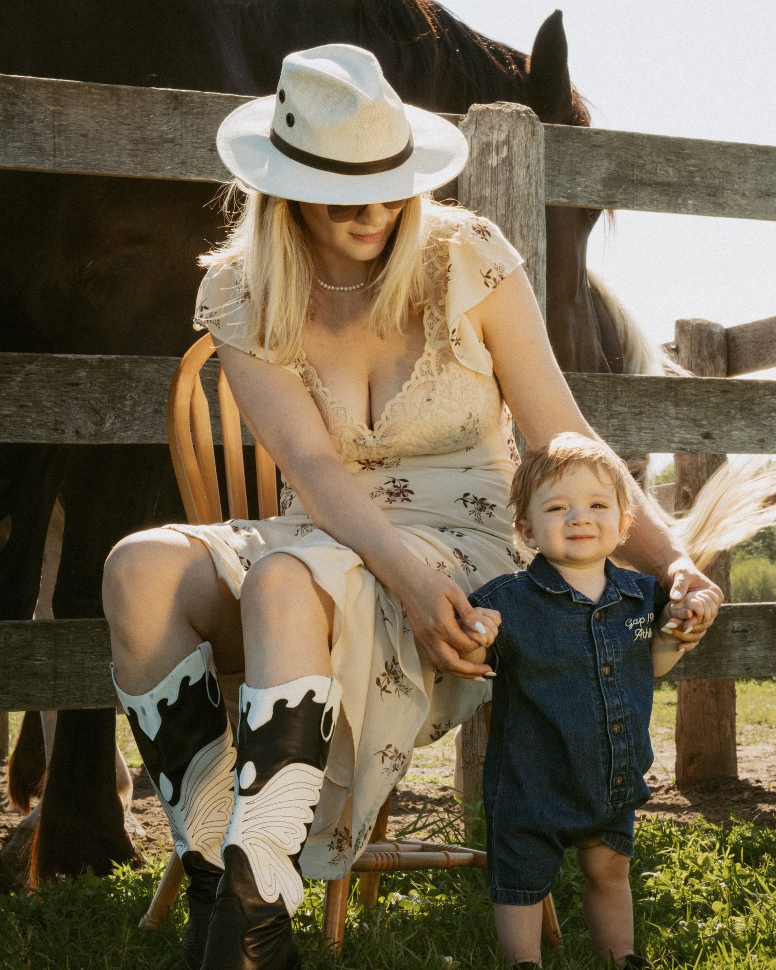 A woman in a floral dress and cowboy boots standing at a paddock fence with her son on her hip