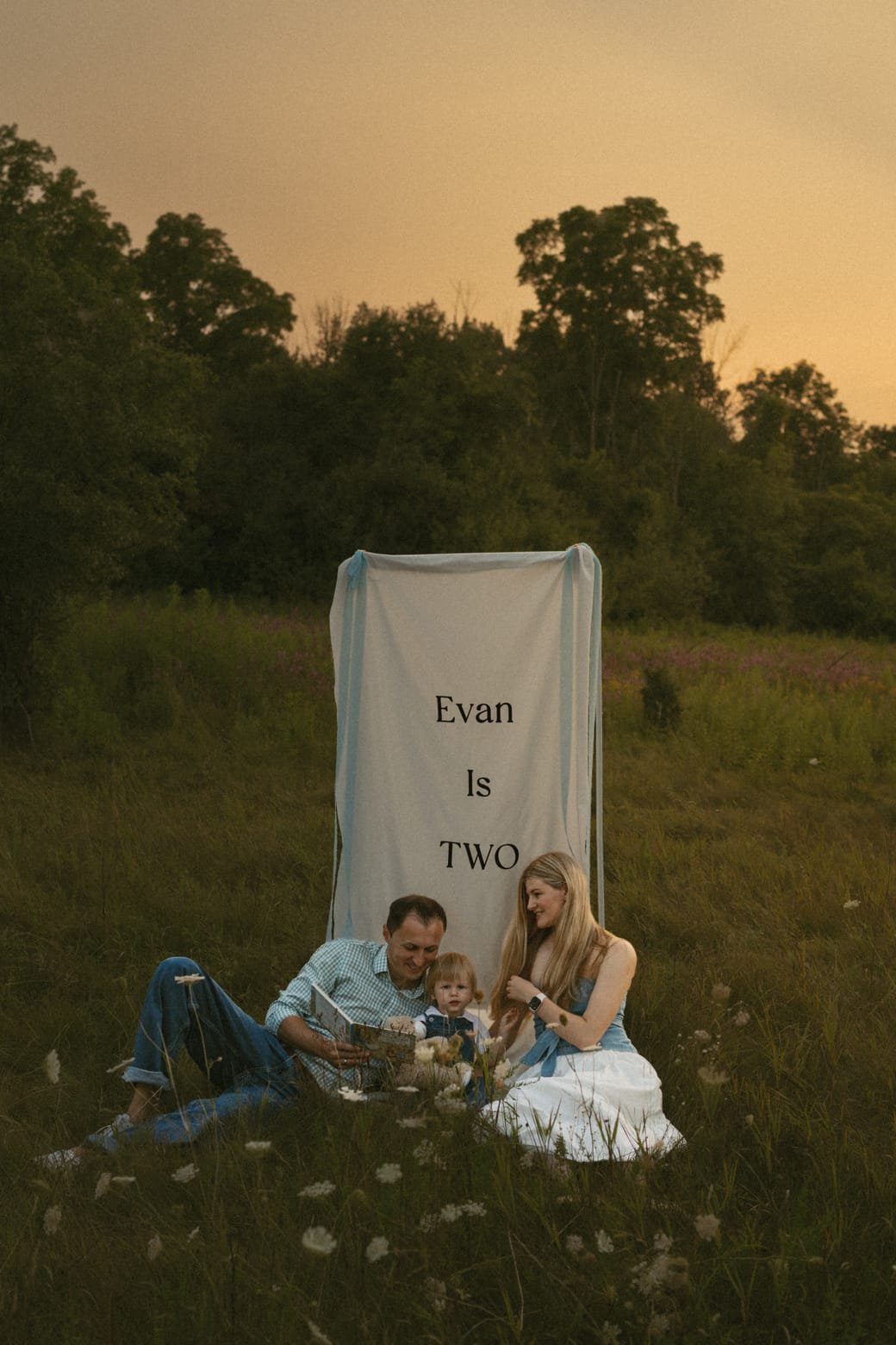 A family of three seated on the grass in front of a linen banner reading Evan Is Two