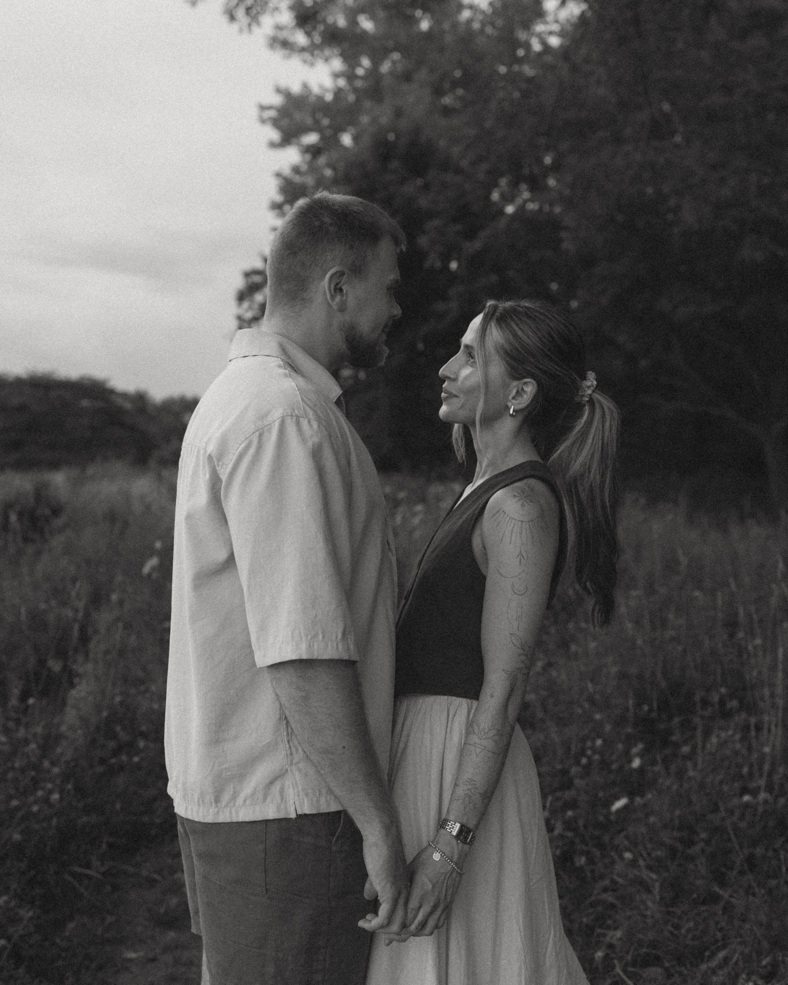 A black and white frame of a couple face to face holding hands beneath dark trees by the lake
