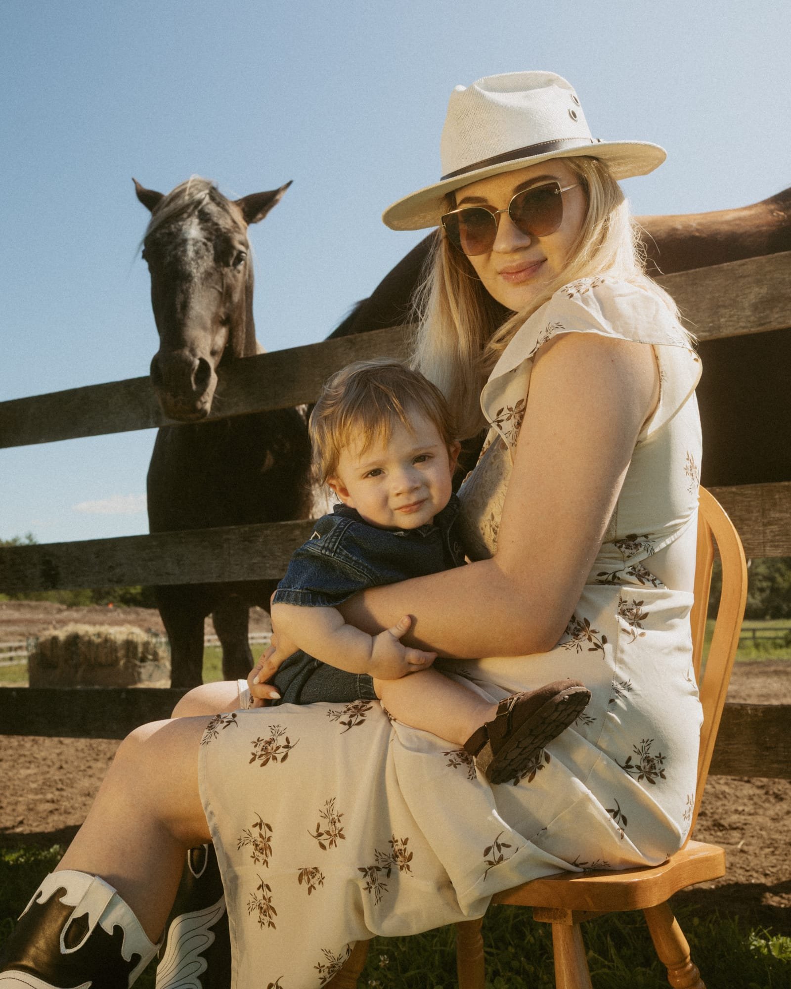 A mother in a wide-brim hat holding her toddler son on her lap beside a horse paddock