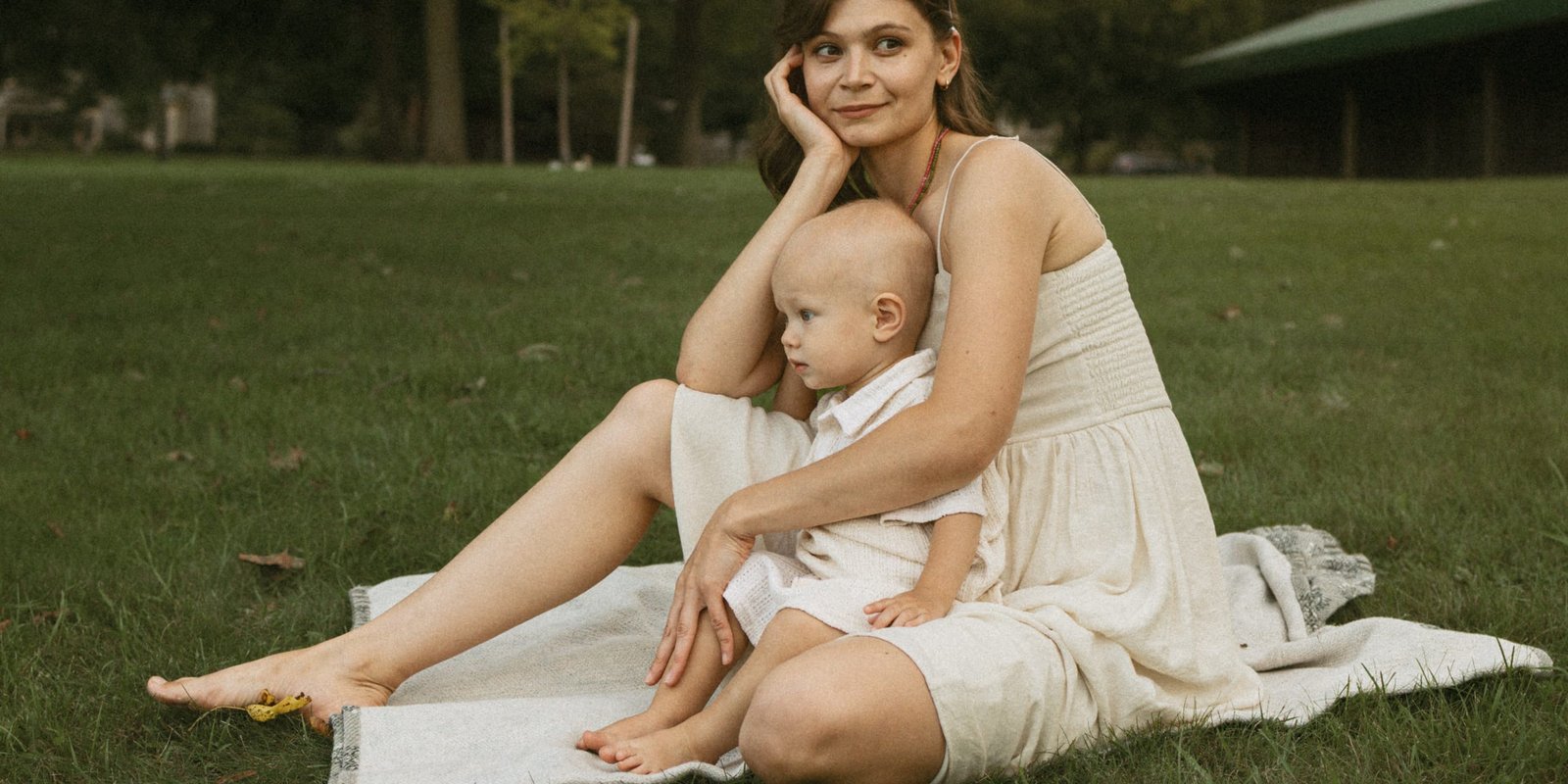 A mother in a cream linen dress sitting barefoot on a blanket in the park with her son in her lap
