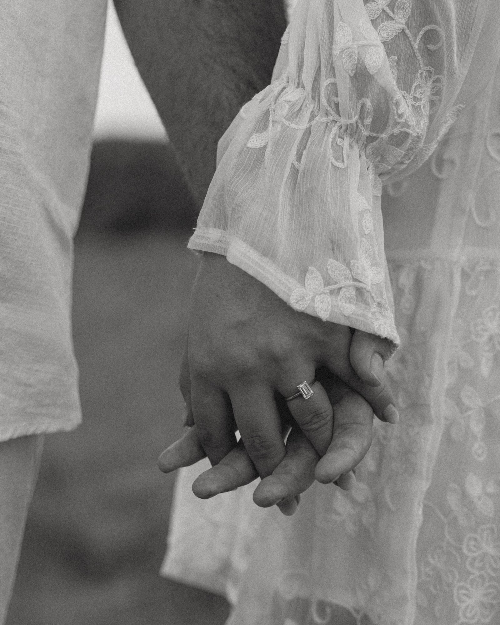 A black and white detail of a couple's hands clasped together against a lace dress